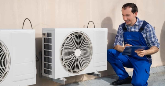 White man in blue overalls kneeling next to a ventilation system writing on a clipboard