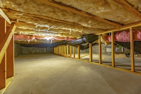 Basement or crawl space with upper floor insulation and wooden support beams. An area of limited height under the floor of a house with concrete wall and floor.