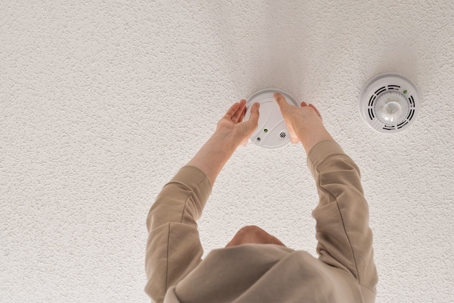 Woman reaching up for smoke alarm on ceiling at home, changing battery time, installation or regular maintenance concept
